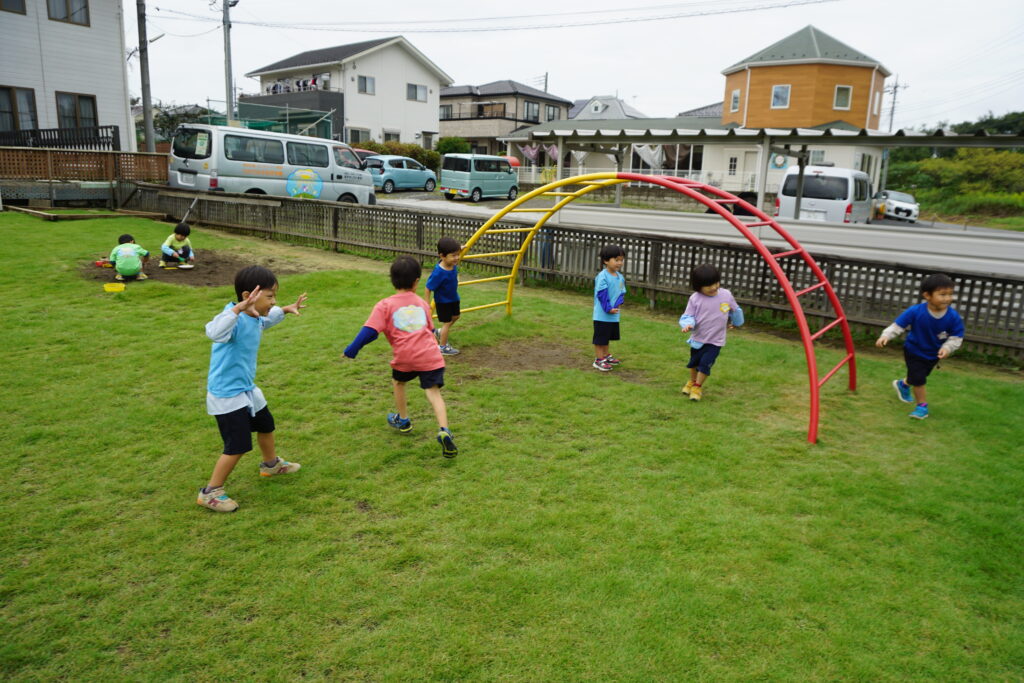 園庭の雲梯の前で鬼ごっこをする子供達とその奥で砂遊びをする子供