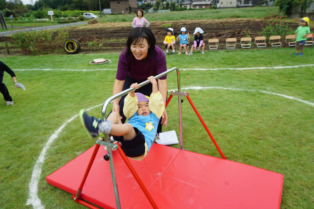 園庭で赤い鉄棒にぶらさがる園児とそれを支える先生