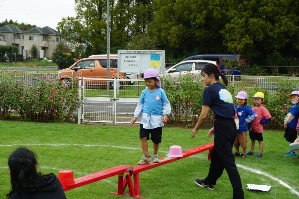 赤い平均台を慎重に渡る男の子とそれを見守る先生達と順番を待つ子供達