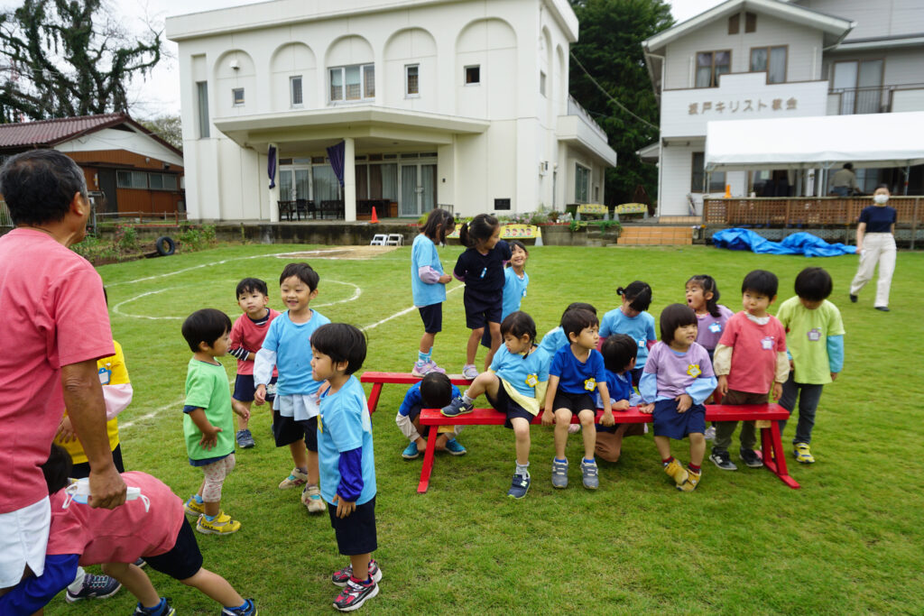 園庭に集まって椅子にならんで座るたくさんの子どもたちとその前で子供と遊ぶ園長